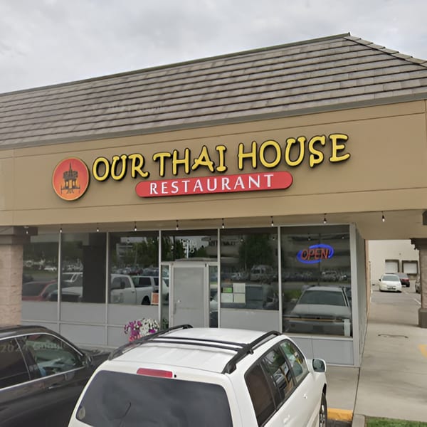 Exterior of Our Thai House restaurant with a large sign and glass windows, cars parked outside.