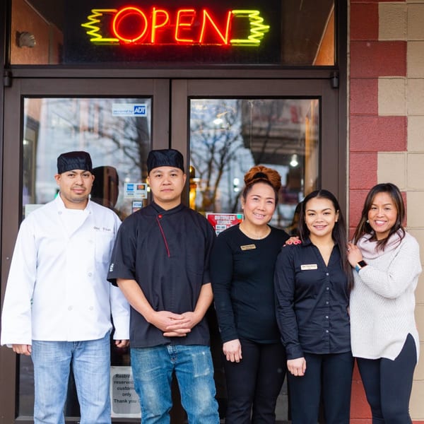 Five Our Thai House staff members, including chefs and servers, smiling in front of the restaurant entrance.