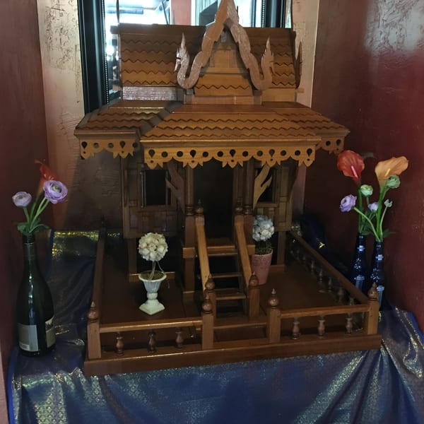 A wooden Thai spirit house with flowers in vases, set against a red wall and blue fabric.
