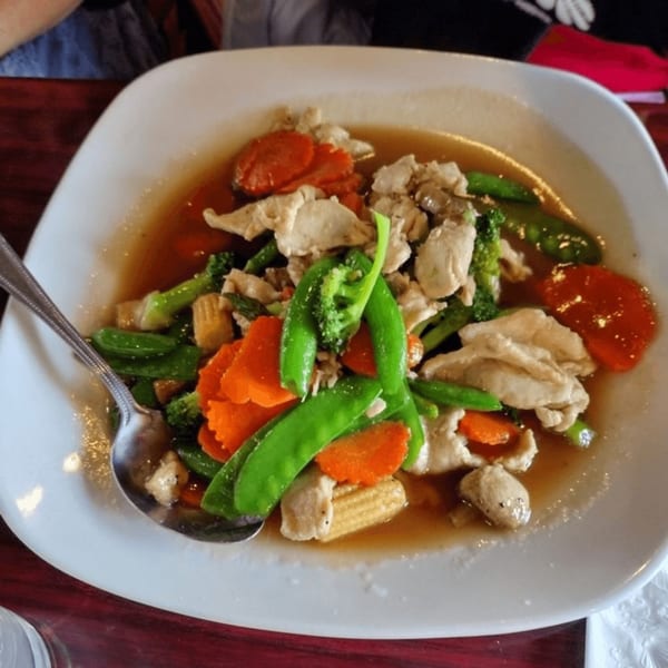 A Thai dish with chicken, broccoli, carrots, and snow peas in a savory sauce on a white plate.
